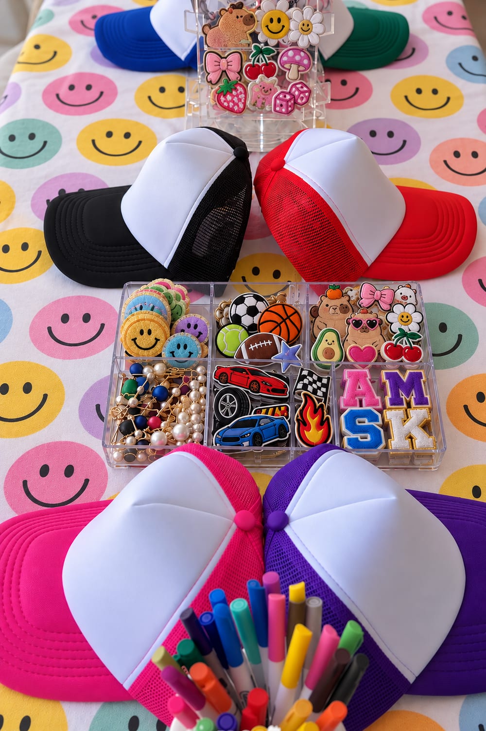 Trucker caps in zwart, rood, roze en paars op een smiley-tafelkleed, met een bak vol patches en stiften